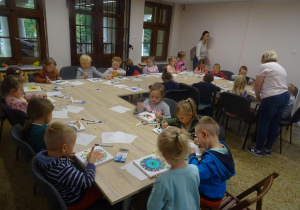 Grupa dzieci przy dużym stole maluje na płótnie. Nad stołem pochylona jest pani Agnieszka, która pomaga dzieciom podczas pracy.